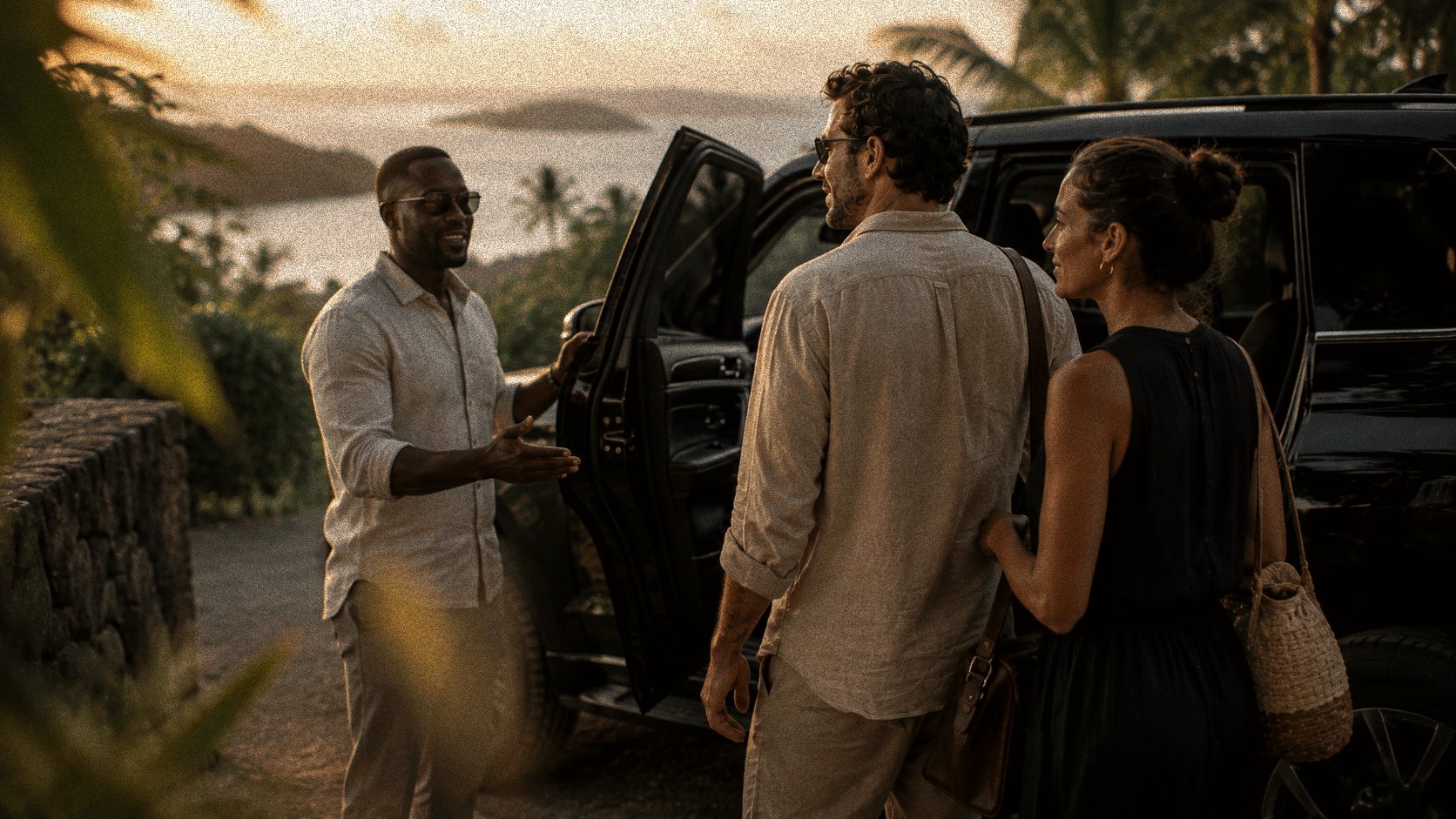 A host welcoming guests and opening a car door at golden hour, sea and islands beyond